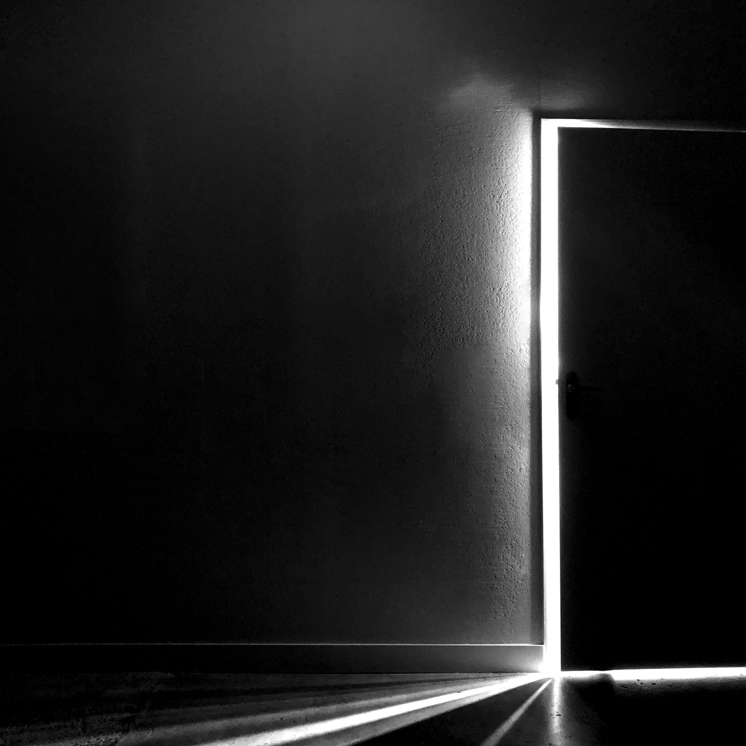 Photographie en noir et blanc d'un porte très légèrement entre ouverte laissant passer un filet de lumière dans une pièce sombre. L'encadrement de la porte s'arrête au tiers de la photo, qui est en format carré.